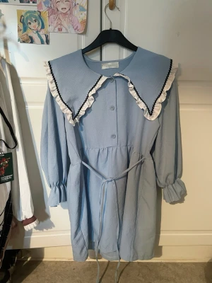 Ljusblå klänning med volangkrage - Supersöt ljusblå klänning från dream shop(?) med stor volangkrage dekorerad med vit och svart kant. Klänningen har knappar framtill, långa puffärmar med resår vid ärmslut och ett knytband i midjan. Materialet är lätt och har en strukturerad yta. Köpt utomlands för flera år sedan, så jag minns inte priset men jag tror runt 169kr. Säljer för 90kr. Använd fåtal gånger utan defekter