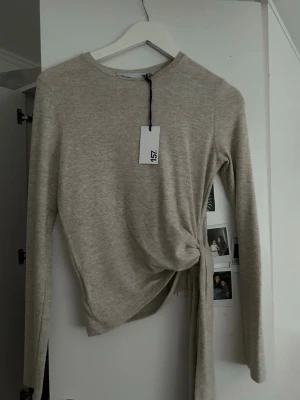 Beige långärmad topp med knyt - Trendig beige långärmad topp från 157 med rund halsringning och snygg knytdetalj i sidan. Tillverkad i mjukt material som känns skönt mot huden. Perfekt att styla med jeans eller kjol för en clean look.