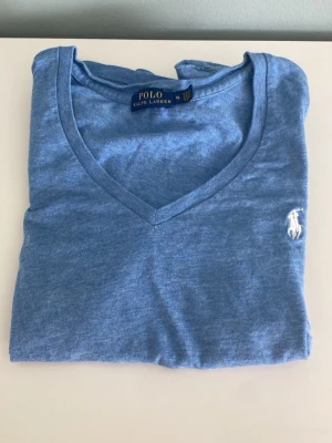 Blå v-ringad t-shirt från Polo Ralph Lauren - Snygg blå t-shirt från Polo Ralph Lauren med v-ringning och klassisk vit logga broderad på bröstet. Tillverkad i mjuk bomull som känns skön mot huden. Perfekt till jeans eller shorts för en clean och enkel stil.