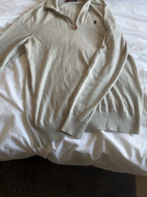 Beige stickad tröja från Morfils - Snygg beige stickad tröja från Morfils i 100% merinoull. Tröjan har halv dragkedja vid halsen, ribbade muddar och en liten broderad logga på bröstet. Perfekt för lager på lager och enkel att matcha med jeans eller chinos.