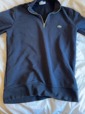 Marinblå hoodie från Lacoste - Snygg marinblå hoodie från Lacoste med halv dragkedja och klassisk krokodillogga på bröstet. Tillverkad i mjukt bomullsmaterial och har ribbade muddar. Perfekt för en chill och stilren look.