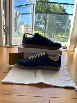 Blå sneakers Maison Margiela - Snygga blå sneakers från Maison Margiela med beige snörning och gummisula. Skorna har en mix av skinn och mocka i ovandelen och klassisk låg profil. Perfekta för dig som gillar stilrena och trendiga sneakers.