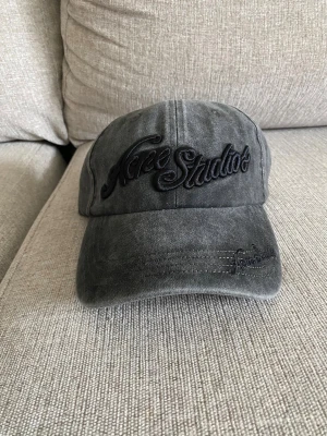 Grå keps från Acne Studios - Säljer en grå keps från Acne Studios med broderad logga framtill i svart. Kepsen har böjd skärm och justerbart spänne bak. Tillverkad i tvättad bomull för en avslappnad look.