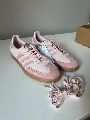 Adidas Samba rosa sneakers - Snygga Adidas Samba sneakers i ljusrosa och beige mocka med klassiska tre ränder på sidan och gummisula. Skorna har lågt skaft och kommer med extra skosnören. Perfekta för dig som vill ha en stilren och trendig look.