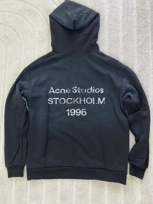 Svart hoodie Acne Studios Stockholm - Svart hoodie från Acne Studios med stor huva och tryck på ryggen där det står 'Acne Studios STOCKHOLM 1996'. Klassisk passform med ribbade muddar och mjukt material, perfekt för en clean och stilren look.