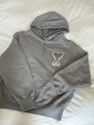 Grå Ami Paris hoodie - Säljer en stilren grå hoodie från Ami Paris med stor vit broderad logga. Perfekt för en avslappnad och trendig stil. Pris är förhandlingsbart.
