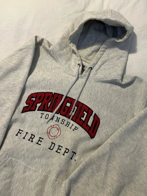 Grå vintage hoodie - Ljusgrå hoodie med huva och dragsnören, stor magficka och tryck. Perfekt för dig som gillar college-vibe och vintage stil.