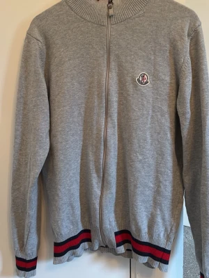 Grå ziptröja från Moncler - Snygg grå ziptröja från Moncler med ribbad krage och muddar. Tröjan har hel dragkedja, broderad Moncler-logga på bröstet och detaljer i rött, blått och svart vid ärmslut och nederkant. Perfekt till jeans eller chinos för en clean look. Måtten från axel söm till axelsöm 40 cm och längd 66cm. Det står L på lappen men det är mer en S/M