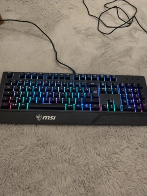 MSI RGB-tangentbord & gamingmus - Säljer ett MSI tangentbord med snygg RGB-belysning och ett gaminginriktat utseende. Tangentbordet är svart med regnbågsfärgade LED-lampor. Medföljer även en svart gamingmus med coolt mönster och ergonomisk form, perfekt för gaming eller vardagsbruk.