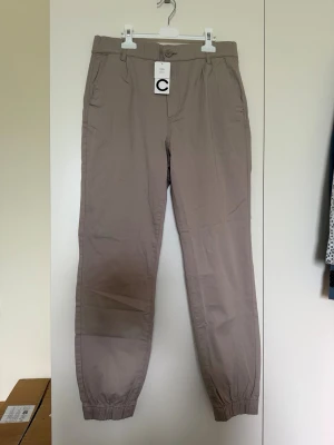 Beige mjukisbyxor från C - Snygga beige mjukisbyxor från Cubus med elastiska benslut och klassisk passform. Byxorna har fickor på sidorna och är tillverkade i ett mjukt och lätt material bomull som är perfekt för chill dagar. Stängs med knapp och dragkedja framtill.Helt nya