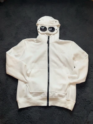 Vit hoodie med glasögon från C.P. Company - Säljer en vit hoodie från C.P. Company med hel dragkedja och ikoniska glasögon i huvan. Hoodien har två stora fickor framtill och är tillverkad i mjukt bomullsmaterial. Perfekt för dig som vill sticka ut med en unik detalj på huvan.