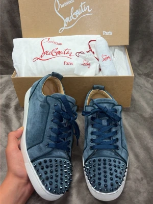 Washed blue Christian Louboutin sneakers - Säljer ett par blåa sneakers från Christian Louboutin i mocka med matchande blå skosnören och vita sulor. Skorna har coola nitar på tån som ger en edgy vibe. Perfekta för dig som vill sticka ut med din stil.