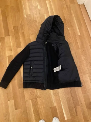 Marinblå Moncler pufferjacka med huva - Marinblå pufferjacka från Moncler (äkta) med stickade ärmar och detaljerad ficka med dragkedja på ena ärmen. Jackan har huva som är bekväm och som är allmänt väldigt snygg, den är i storlek S men passar även i XS. Jackan är använd ett få tal gånger och är nästan som helt ny.