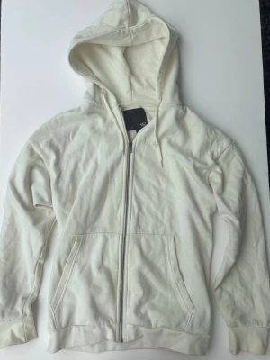 Vit zip hoodie från 157 - Klassisk vit hoodie från 157 med dragkedja framtill och två stora fickor. Hoodien har justerbar huva med snören och är tillverkad i mjukt bomullsmaterial. Perfekt för en avslappnad och enkel stil.