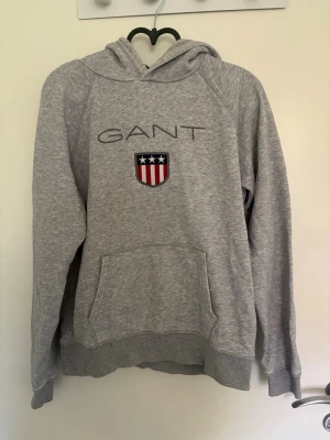 Grå hoodie från GANT med logga - Snygg grå hoodie från GANT med broderad logga och amerikanskinspirerat emblem på bröstet. Klassisk känguruficka framtill och huva. Perfekt för en avslappnad och sportig stil. Tillverkad i mjukt sweatshirtmaterial.