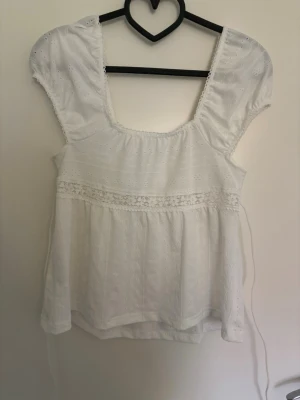 Vit blus med broderade detaljer - Söt vit blus med broderade detaljer och puffärmar. Toppen har fyrkantig ringning, knytband i midjan och spetsinslag. Perfekt för en somrig look och passar till både jeans och kjol.