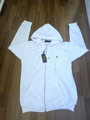 Vit hoodie med dragkedja från Polo Ralph Lauren - Säljer en clean vit hoodie från Polo Ralph Lauren med dragkedja framtill, huva med dragsko och två stora fickor. Klassisk liten logga broderad på bröstet. Perfekt för en chill och stilren look. Enda defekten är i polo tag där typ 2 bokstäver har fallit av