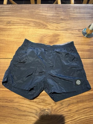Mörkblå badbyxor från Stone Island - Snygga mörkblå badbyxor från Stone Island med elastisk midja och sidofickor. Klassisk kort modell med logotyp-patch på vänster ben. Materialet är lätt och snabbtorkande, perfekt för stranden eller poolen.
