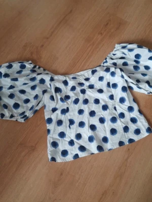 Vit topp med blå prickar och puffärm - Trendig vit topp med stora blå prickar och rejäla puffärmar. Offshoulder-modell som ger en cool och lekfull vibe. Perfekt för dig som vill sticka ut lite extra. Tillverkad i ett lätt bomullstyg som känns skönt mot huden.