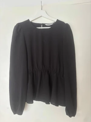 Svart blus från ONLY med rynkad midja - Svart blus från ONLY med rund halsringning och rynkad midja som ger en snygg siluett. Blusen har långa ärmar med lätt puff och resår vid ärmslut. Tillverkad i ett tunt, flowy tyg som ger ett stilrent intryck.