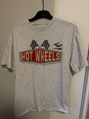 Grå Hot Wheels t-shirt från H&M - Säljer en grå t-shirt från H&M:s DIVIDED-kollektion med stort Hot Wheels-tryck framtill och coola rutiga flaggor. T-shirten har rund hals och korta ärmar. Perfekt för dig som gillar streetwear och retro vibes.