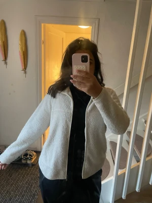 Vit fleece jacka med dragkedja - Mysig vit cardiganjacka i fleece med hel dragkedja framtill och hög krage. Jackan har en avslappnad passform och långa ärmar, perfekt för lager på lager under kyliga dagar. Enkel och stilren design som passar till det mesta.