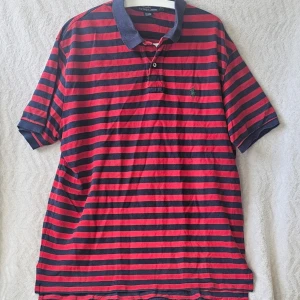 Randig pikétröja från Polo Ralph Lauren - Klassisk pikétröja från Polo Ralph Lauren i rött och marinblått med horisontella ränder. Tröjan har krage, knappar vid halsen och den ikoniska broderade loggan på bröstet. Perfekt till jeans eller shorts för en avslappnad look.