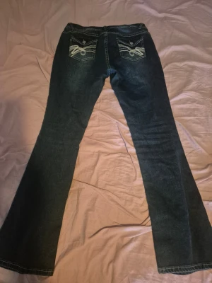 Blå bootcut jeans med broderade fickor - Säljer ett par blå bootcut jeans med snygga broderade detaljer på bakfickorna och kontrastsömmar. Jeansen har klassisk femficksdesign och stängs med knapp och dragkedja. Perfekta för dig som gillar en retro och avslappnad stil.