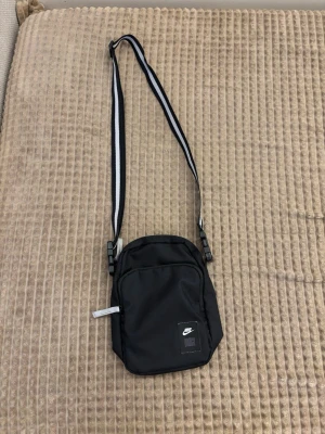 Svart axelväska från Nike - Snygg svart axelväska från Nike med justerbar axelrem i svart och vitt. Väskan har en praktisk framficka med dragkedja och en diskret Nike-logga på framsidan. Tillverkad i slitstarkt syntetmaterial, perfekt för att bära det viktigaste.