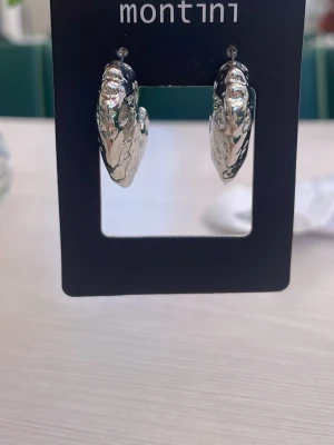 Silvriga chunky örhängen Montini - Säljer ett par chunky örhängen från Montini i blankt silverfärgat metallmaterial. De har en cool, ojämn och organisk form som ger en trendig look. Perfekta för dig som gillar statement-smycken och vill sticka ut lite extra.