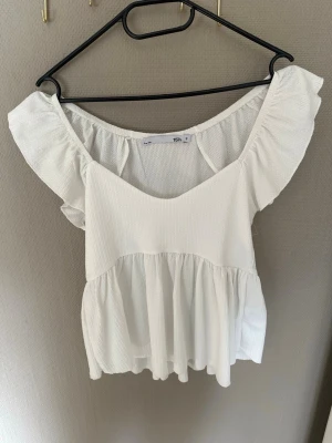 Vit volangblus - Söt vit blus med volangdetaljer. Toppen har en luftig passform och är tillverkad i ett lätt, strukturerat tyg. Perfekt för varma dagar och ger en romantisk vibe till din outfit.