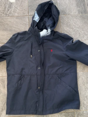 Marinblå vindjacka från Polo Ralph Lauren - Snygg marinblå vindjacka från Polo Ralph Lauren. Luvan går att ta av du stänger jackan via dragkedja eller knappar. Priset går att diskutera