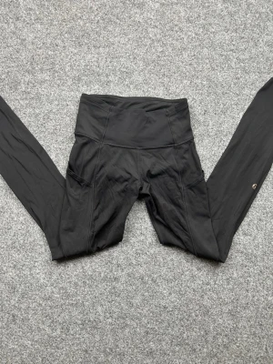Svarta leggings från Lululemon - Snygga svarta leggings från Lululemon med hög midja och praktiska sidofickor. Tillverkade i ett stretchigt och mjukt material som sitter skönt på kroppen. Perfekta för träning eller chill. Liten Lululemon-logga på benet.