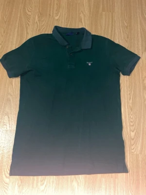 Grön pikétröja från Gant - Klassisk grön pikétröja från Gant med korta ärmar och krage. Tröjan har knappar vid halsen och en liten broderad logga på bröstet. Perfekt till jeans eller shorts för en avslappnad stil.