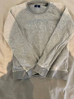 GANT grå tröja (äkta) - Snygg grå sweatshirt från GANT med broderad logga framtill. Tröjan har rund halsringning, långa ärmar och ribbade muddar vid ärmslut och nederkant. Tillverkad i mjukt material som känns skönt mot huden. Perfekt för en avslappnad och stilren look. Storlek 158-164. 