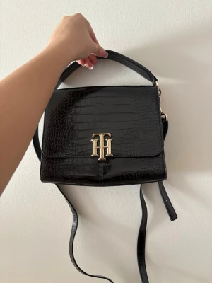 Svart handväska Tommy Hilfiger - Snygg svart handväska från Tommy Hilfiger i croco-mönstrat konstläder. Väskan har guldfärgade detaljer och ett stort TH-logo-spänne framtill. Justerbar axelrem och ett stort topphandtag gör den enkel att bära. Nypris: 2250kr, dustbag medföljer 