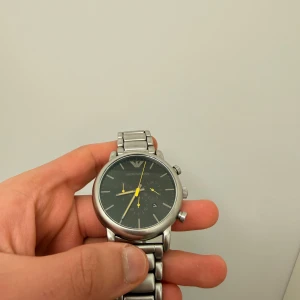 Emporio Armani silverfärgad klocka - Stilren klocka från Emporio Armani med silverfärgad länk i metall och svart urtavla. Klockan har gula visare och kronograf-funktion. Klassisk rund form och diskret logga vid tolvslaget. Perfekt accessoar för en modern look.