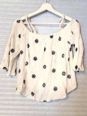 Vit offshoulder blus med broderi - Snygg vit offshoulder blus med svarta broderade blommor och trekvartsärm. Blusen har resår vid axlarna och tunna axelband för extra stöd. Perfekt för en avslappnad och trendig look.