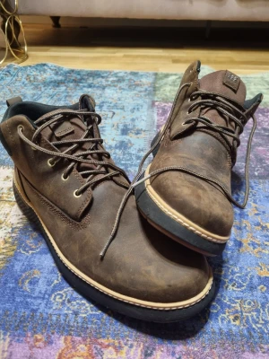 Bruna Timberland kängor i läder - Säljer ett par bruna kängor från Timberland i robust läder med svarta detaljer och grov sula. Skorna har metallöljetter, bruna snören och en vadderad kant runt ankeln för extra komfort. Perfekta för dig som vill ha en klassisk och stilren look. Helt ny aldrig använd.                             Ny pris 2045kr från zalando