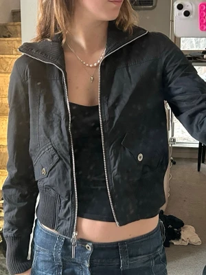 Svart bomberjacka med dragkedja - Snygg svart bomberjacka med silvrig dragkedja och ribbade muddar. Jackan har två fickor framtill med knappstängning och en klassisk kort passform. Perfekt till jeans och crop top för en cool streetstyle-look.