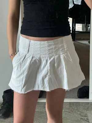 Vit kortkjol med veckad design - Snygg vit kortkjol med veckad design och hög midja. Kjolen har en lätt utsvängd form och råa kanter nertill för en trendig look. Perfekt att matcha med en crop top eller hoodie. Materialet känns som bomull och är skönt mot huden.