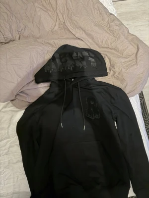 Svart hoodie från Risky med tryck - Svart hoodie från Risky med dragsko i huvan och ett unikt svart tryck på huvan samt en broderad detalj på bröstet. Hoodien har klassisk känguruficka och är gjord i mjukt bomullsmaterial. Perfekt för en streetwear-look.