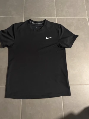 Svart Nike Dri-Fit tränings-tshirt - Säljer en svart tränings-tshirt från Nike med klassisk vit swoosh-logga på bröstet. Tillverkad i lätt och snabbtorkande Dri-Fit-material som andas, perfekt för gymmet eller löprundan. Tidlös design med rund hals och korta ärmar.