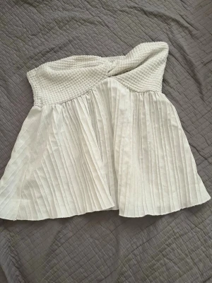Vit bandeau tubtopp med twist - Superfin vit bandeau tubtopp med twistad överdel och plisserad, luftig nederdel. Toppen är axelbandslös och har en söt, flowy passform som passar perfekt till jeans eller kjol. Perfekt för varma dagar och sommarhäng.