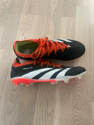 Adidas Predator fotbollsskor svart/vit/röd PRO  - Säljer ett par Adidas Predator fotbollsskor i svart med vita detaljer och röda inslag. Skorna har klassisk snörning, mönstrad ovansida och fasta dobbar för spel på gräs. Snygg design med tydlig Predator-logga undertill. Perfekta för dig som vill sticka ut på planen.