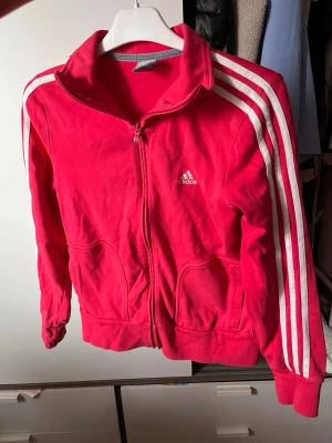 Mörk rosa Adidas zip up  med vita ränder - Snygg mörk rosa hoodie från Adidas med dragkedja framtill och vita ränder längs ärmarna. 