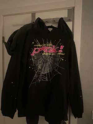 Sp5der Hoodie - Säljer en svart hoodie från Sp5der! med stort färgglatt tryck och spindelnät på framsidan samt en gul stjärna på ryggen. Hoodien har huva och ribbade muddar. Materialet är mjukt och skönt, perfekt för dig som gillar streetwear och unika prints.