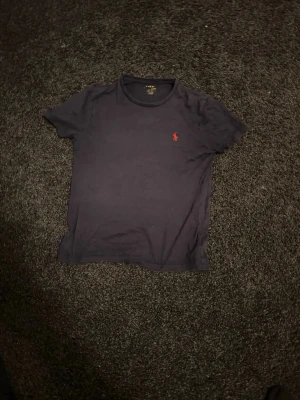 Marinblå t-shirt från Ralph Lauren - Klassisk marinblå t-shirt från Ralph Lauren med den ikoniska röda logotypen broderad på bröstet. T-shirten har rund halsringning och korta ärmar. Tillverkad i mjuk bomull för en skön känsla och enkel stil.