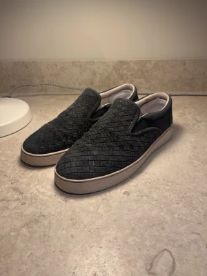 Svarta slip-on sneakers med flätad yta - Säljer ett par svarta slip-on botega veneta sneakers med snygg flätad textur på ovandelen och vit sula. Köpt i butik säljs inte längre i butik så svåra att få tag på. Materialet är mocka. Köpta för 5300 men säljer nu för 2399. Dem är storlek 41.