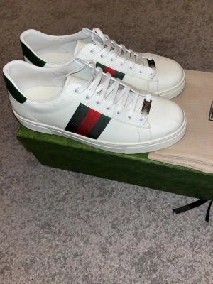 Vita Gucci Ace sneakers - Säljer ett par klassiska vita Gucci Ace sneakers i läder med den ikoniska gröna och röda webbranden på sidan. Skorna har vita snören, metallplatta med Gucci-logga på tån och grön detalj på hälen. Perfekta skor inför vår och sommar.